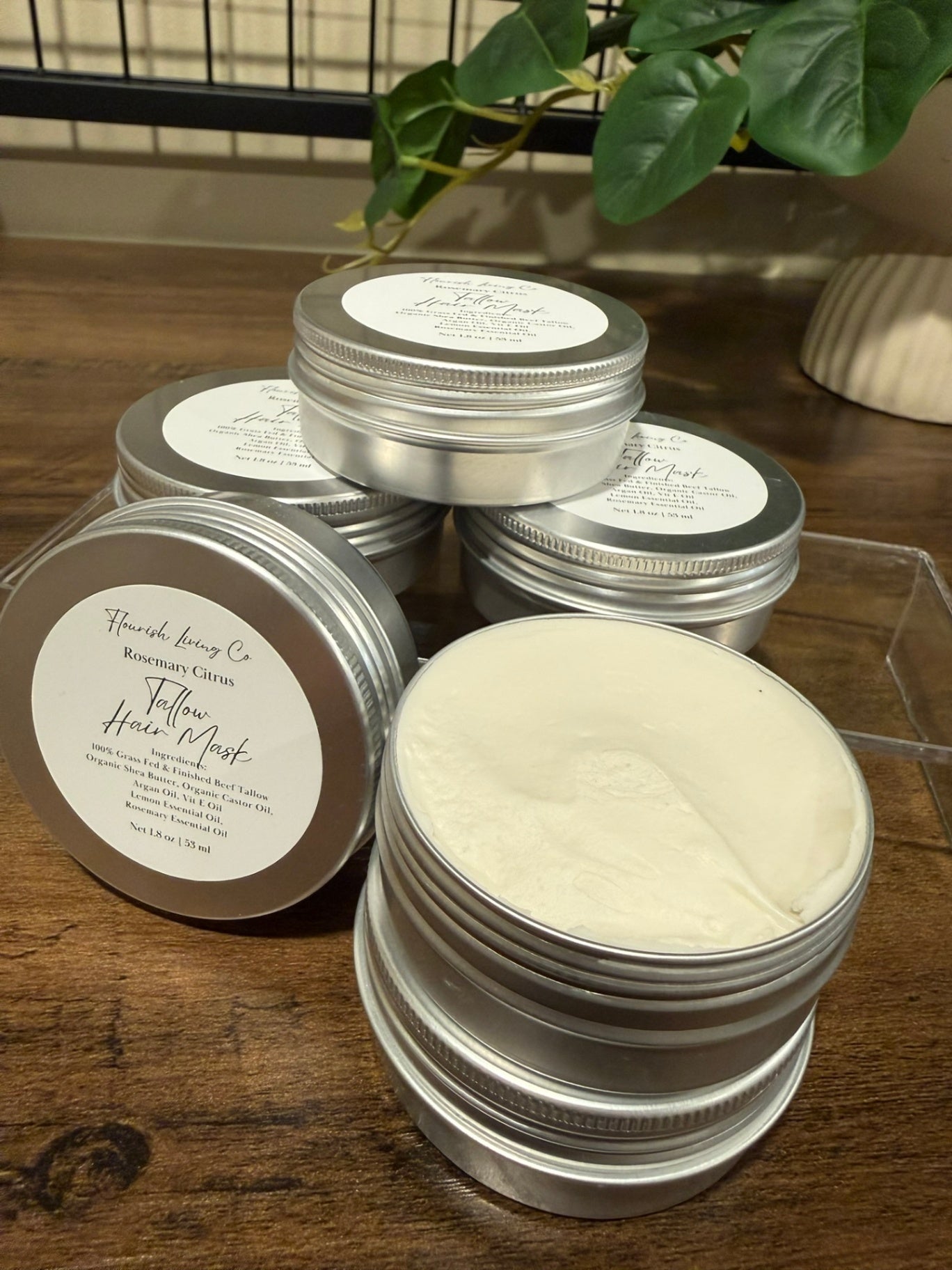 Several round silver metal tins of Flourish Living Co. Tallow Hair Mask are arranged on a wooden tabletop, with one tin open to show the off-white cream product inside and another tin showing the product label.