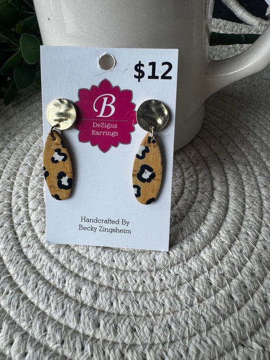 A pair of handmade dangle earrings displayed on a white backing card. The earrings feature a gold-tone flat disc at the top, a tan leather drop with a black leopard print, and a small round gold-tone bead at the bottom.