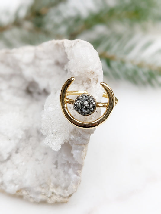 A gold crescent moon ring with a textured black and white druzy stone in the center, displayed on a white crystal cluster with soft green pine branches in the background.
