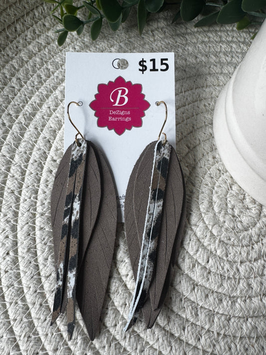 A pair of long feather drop earrings displayed on a white retail card. The earrings feature layered brown feathers with black and white feather accent strips and gold-tone hooks.