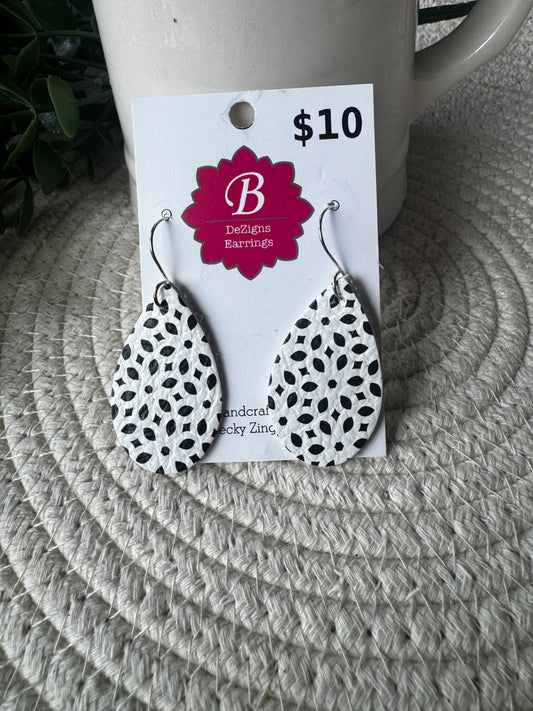A pair of teardrop-shaped dangle earrings with a black and white spotted pattern, attached to a white display card with a magenta flower logo that reads 'B Designs Earrings' and a price of '$10'. The card is resting on a woven circular placemat in front of a white ceramic mug.