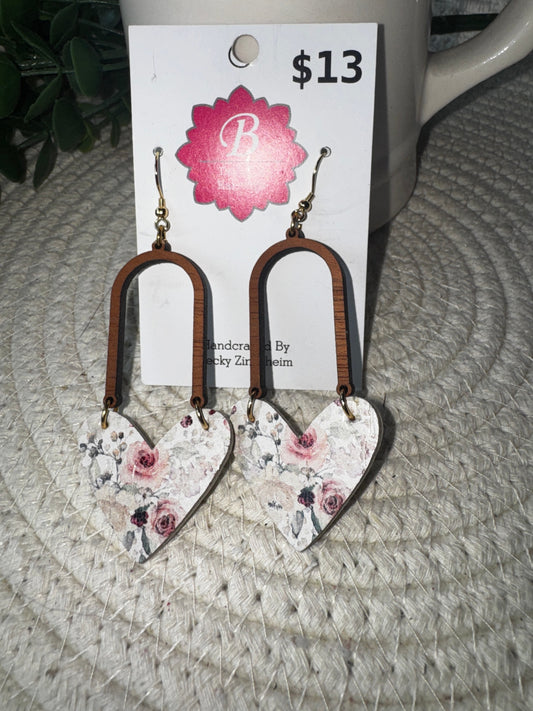 A pair of handmade dangle earrings displayed on a white card labeled '$13' with a pink floral logo. The earrings have brown wooden arches and heart-shaped charms printed with a pink rose floral pattern on a white background, hanging on a woven beige surface.