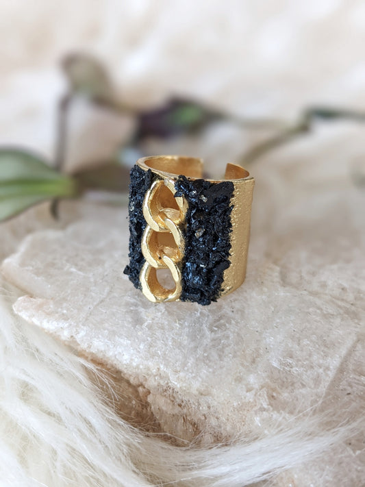 A gold-toned wide band ring with a textured black mineral side and a large interlocking chain-link design on the front, displayed on a soft beige fabric surface.