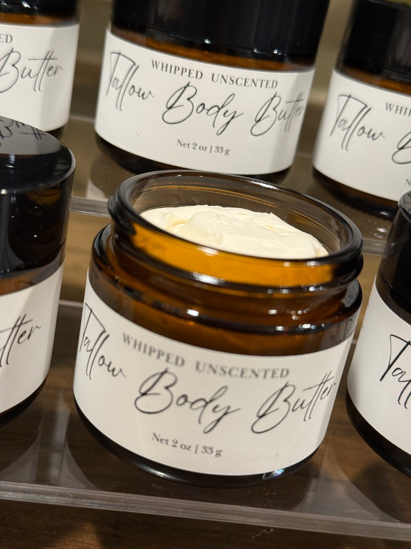 An open amber glass jar of whipped unscented tallow body butter with a white label, displayed on a clear shelf among other matching jars.