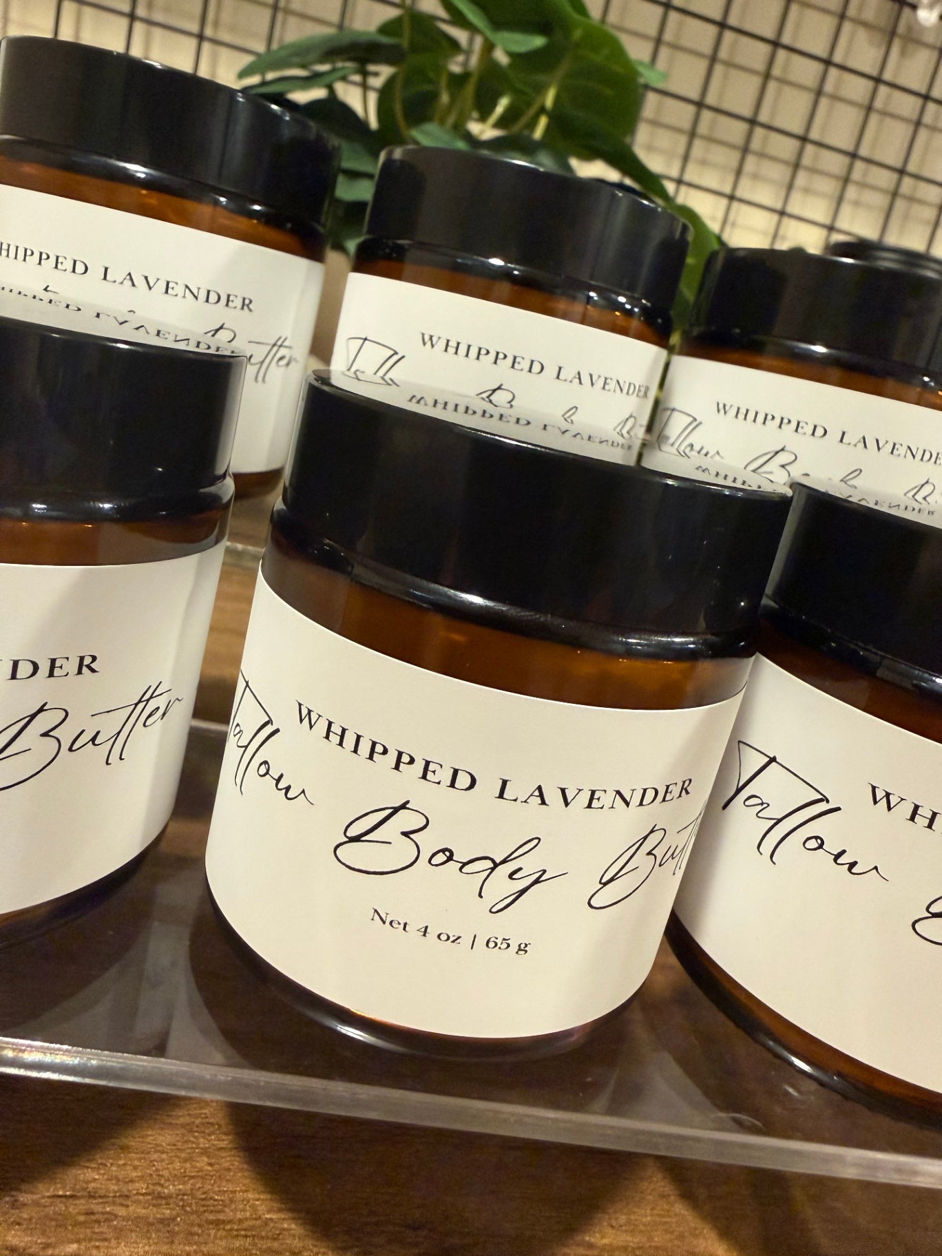 Several amber glass jars of Whipped Lavender Tallow Body Butter with black lids and white labels, arranged on a clear tray on a wooden surface with a plant in the background.