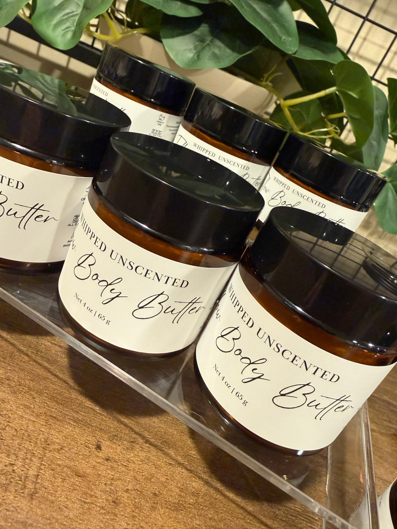 Several amber glass jars of whipped tallow unscented body butter with black lids and white labels, arranged on a clear acrylic shelf over a wooden surface with green leaves in the background.