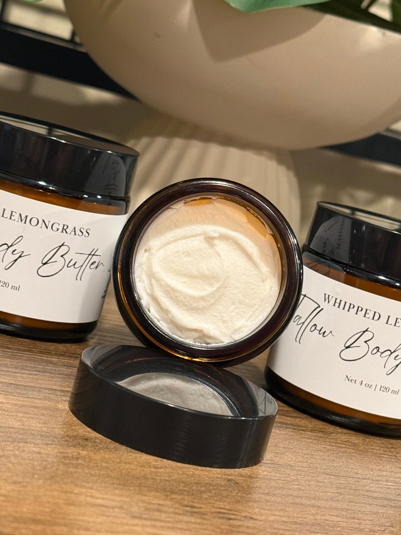 Three amber glass tallow body butter jars with black lids are arranged on a wooden surface. The center jar is open, showing a thick off-white whipped body butter cream inside, while the side jars are closed with white labels featuring black script text.