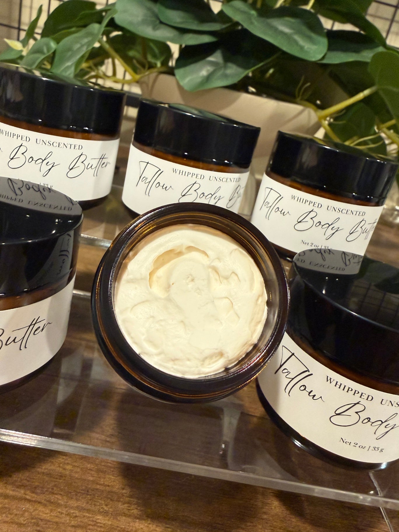 A set of whipped unscented tallow body butter jars with black lids and white labels, with one open jar in the center showing the pale cream-colored body butter. The jars are arranged on a clear acrylic stand on a wooden surface with green plants in the background.