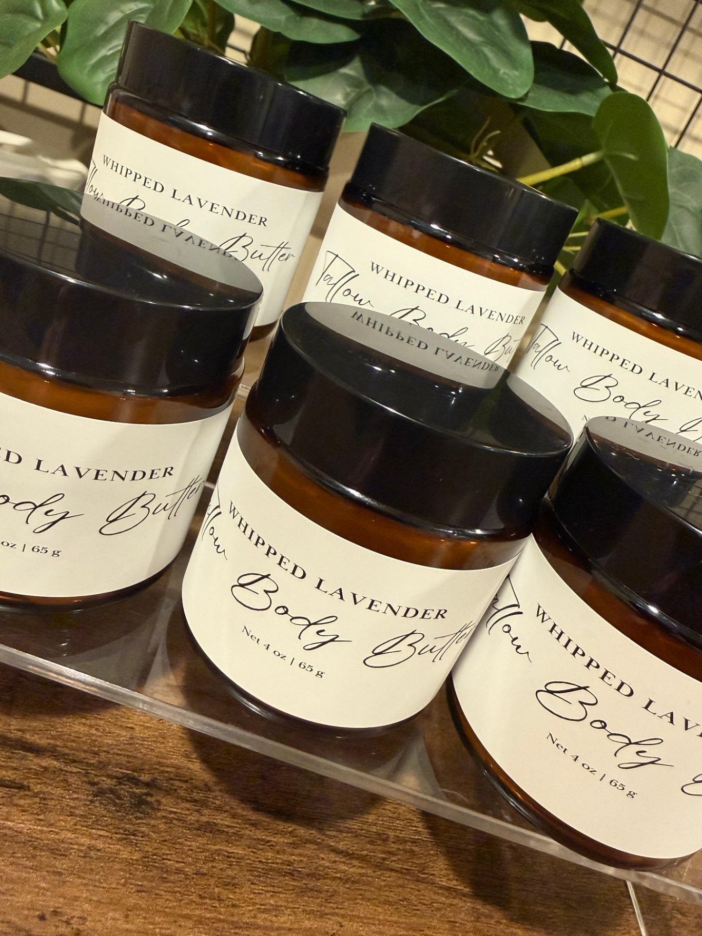 Several amber glass jars of whipped lavender tallow body butter with black lids and white labels reading 'Whipped Tallow Lavender Body Butter' arranged on a clear tray over a wooden surface, with green leaves in the background.