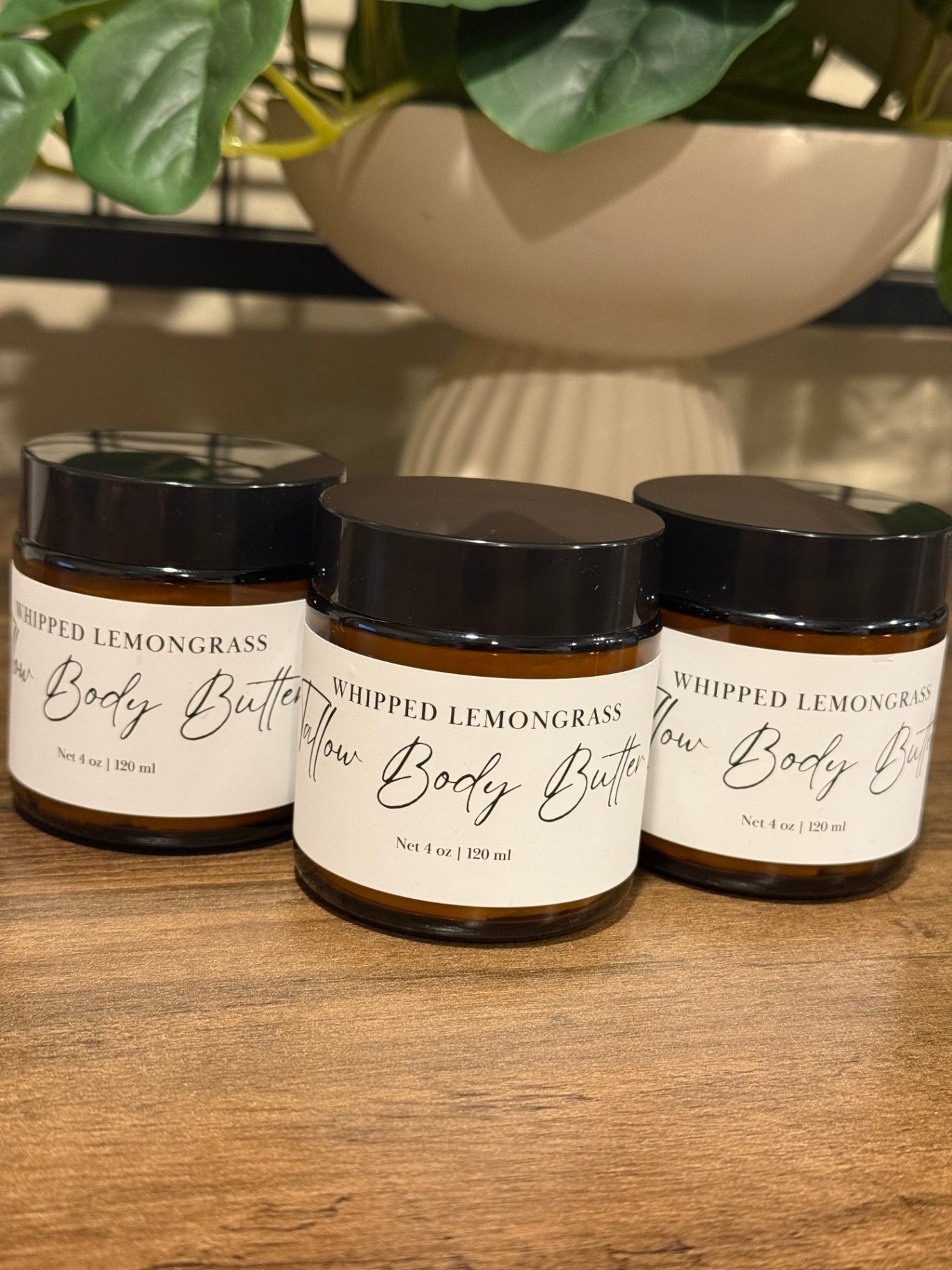 Three amber glass jars of whipped tallow lemongrass body butter with black lids and white labels, arranged on a wooden surface with a potted plant in the background.