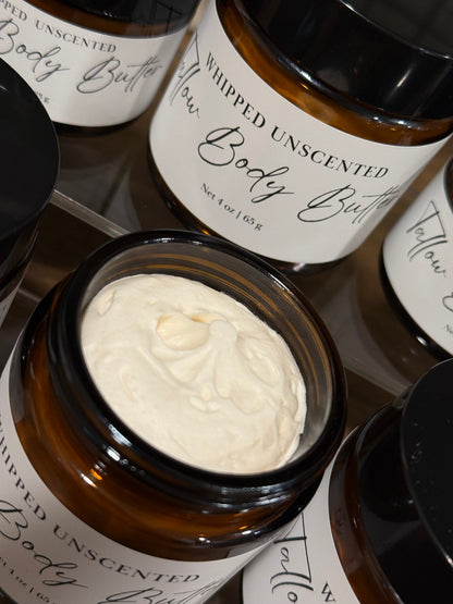 An open amber glass jar of whipped tallow unscented body butter showing the thick off-white cream inside, surrounded by other closed jars with white labels and black lids.