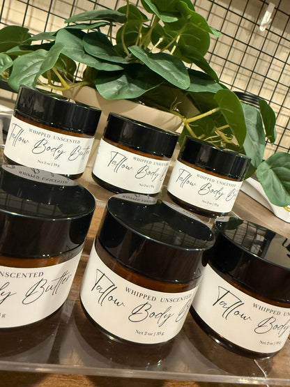Several amber glass jars of whipped Tallow Body Butter with black lids and white labels are arranged on a shelf behind a large green plant, with a tiled wall in the background.