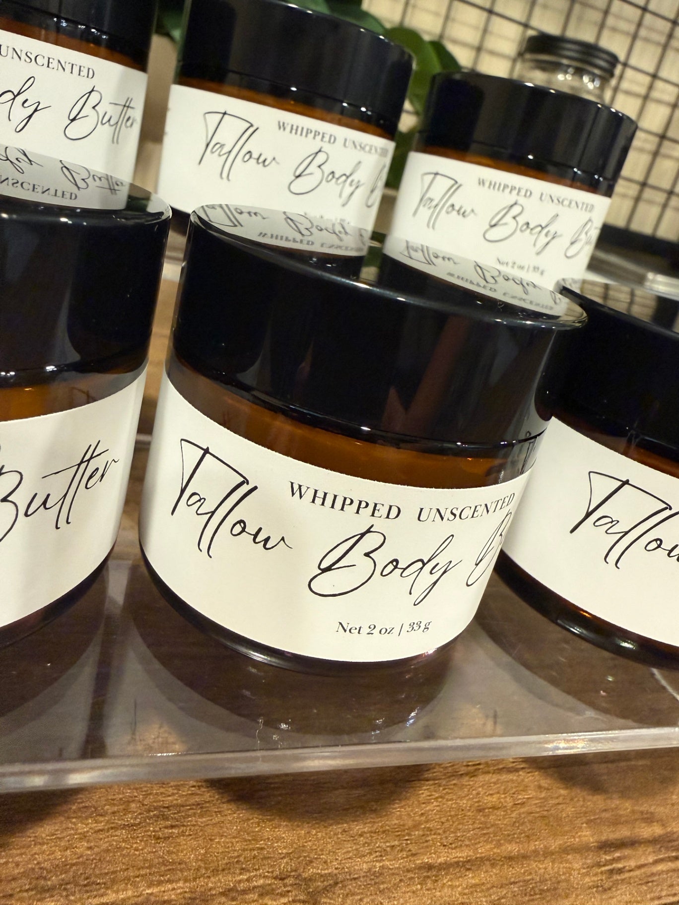 Amber glass jars of whipped tallow body butter with black lids and white labels, displayed on a clear acrylic stand on a wooden surface.
