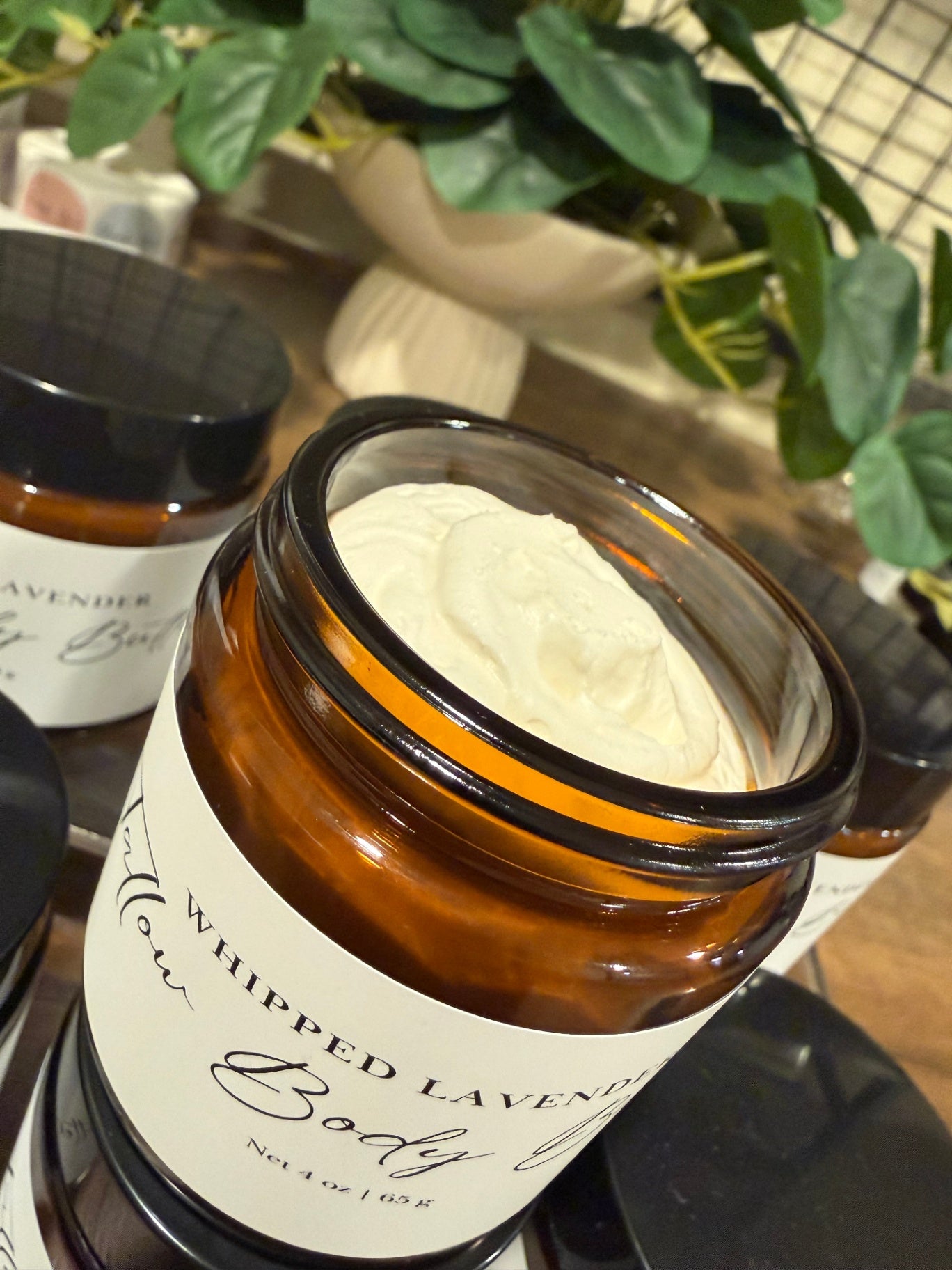 An amber glass jar candle labeled 'Whipped Lavender Body Butter' with white cream visible inside, shown in a styled setting with other jars and green plants in the background.