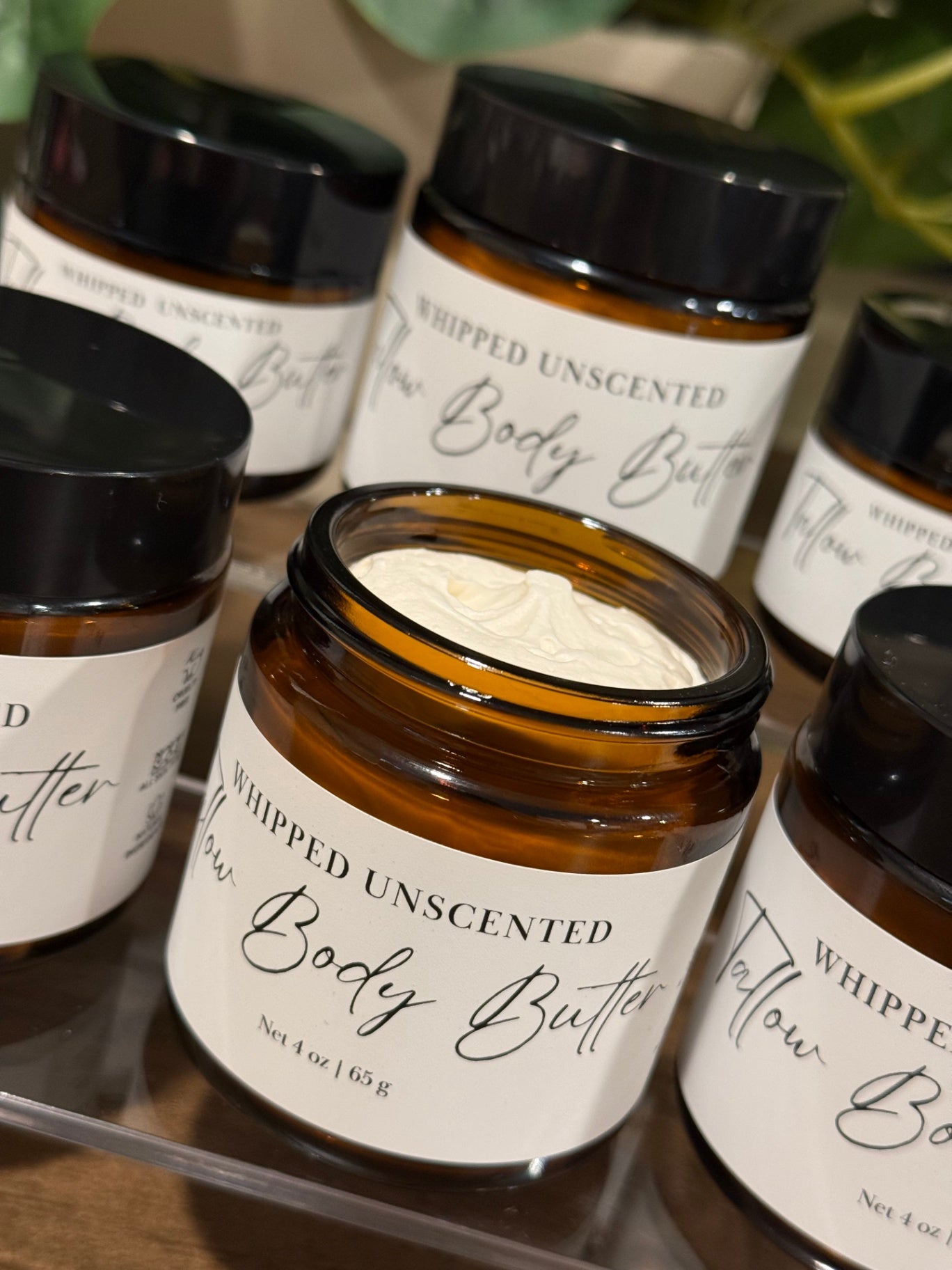 An open amber glass jar of whipped tallow unscented body butter with a white cream inside, surrounded by other closed jars on a clear acrylic tray.
