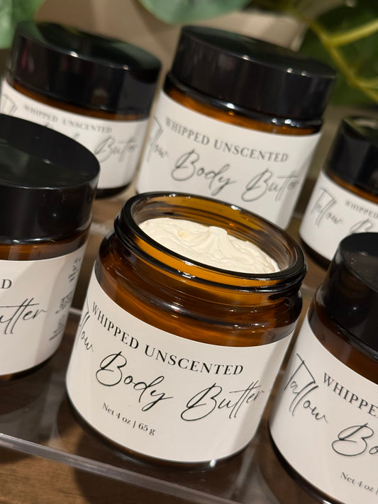 An open amber glass jar of whipped tallow unscented body butter with a white cream inside, surrounded by other closed jars on a clear acrylic tray.