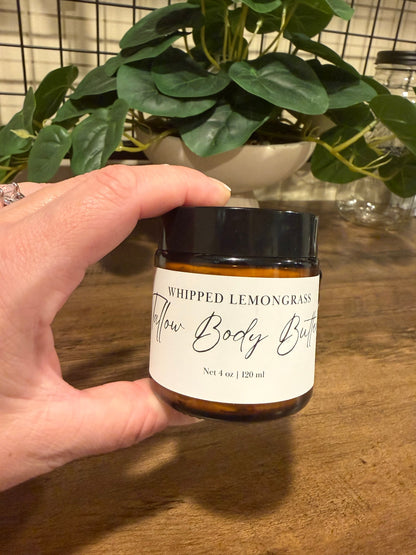 A hand holding a small amber glass jar of whipped tallow lemongrass body butter with a black lid and white label, set against a wooden table with a potted plant in the background.