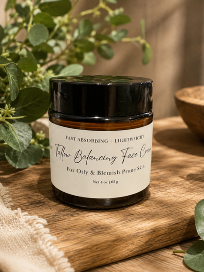 A jar of 'Tallow Balancing Face Cream' in an amber glass jar with a black lid and a white label, styled on a wooden tray with green leaves and a neutral background.