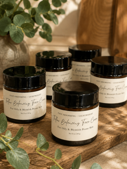Five small black jars of Tallow Balancing Face Cream arranged on a wooden surface with green leaves around them. The front jar is in focus, while the others are softly blurred in the background.