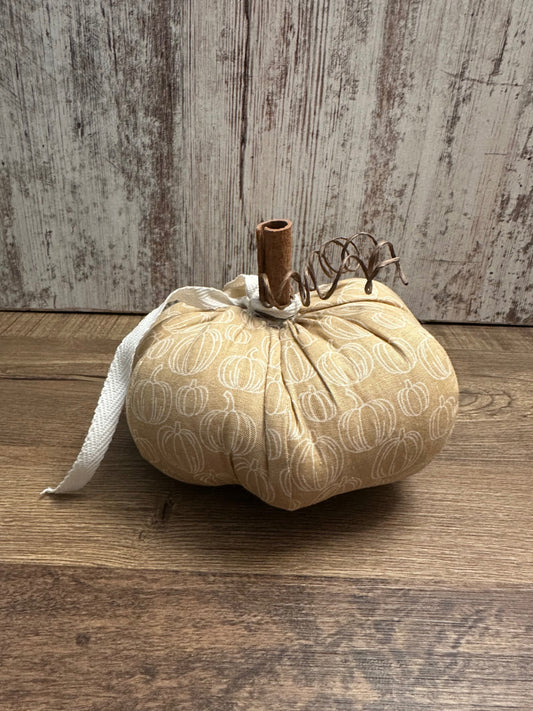 A fabric pumpkin decoration in a warm beige color with a white pumpkin pattern, featuring a cinnamon stick stem and a white ribbon, set on a wooden surface against a rustic wood plank background.