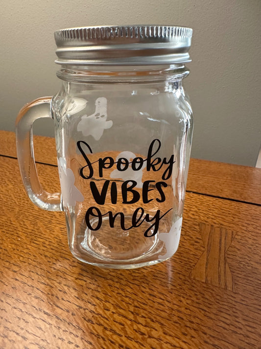 A clear glass mason jar mug with a silver metal lid and handle, featuring the text 'Spooky Vibes Only' in black script and white ghost graphics, sitting on a wooden table.