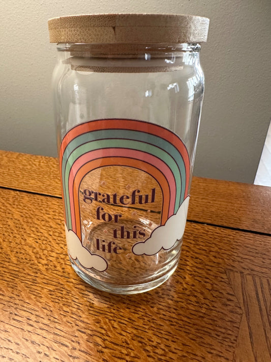A clear glass jar with a natural wood lid, featuring a retro rainbow and cloud graphic with the text 'grateful for this life' on the front, set on a wooden table.