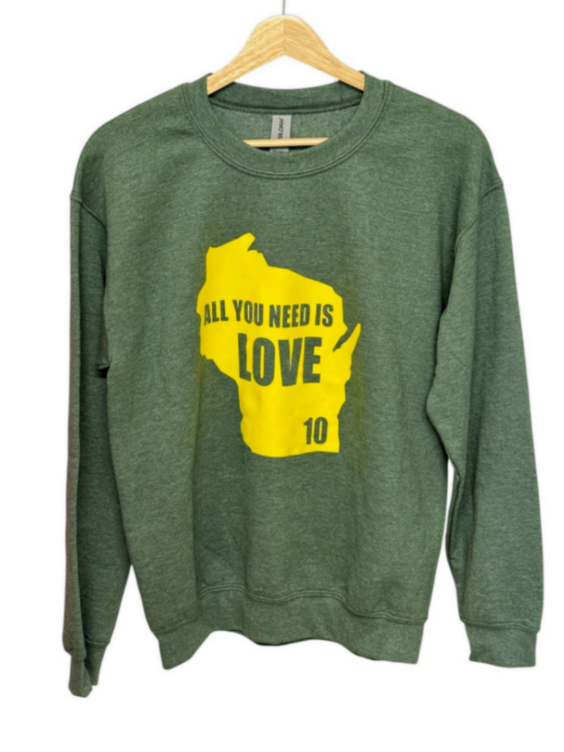 A green crewneck sweatshirt hanging on a wooden hanger against a white background, featuring a yellow graphic of the state of Wisconsin with the text 'ALL YOU NEED IS LOVE 10'.