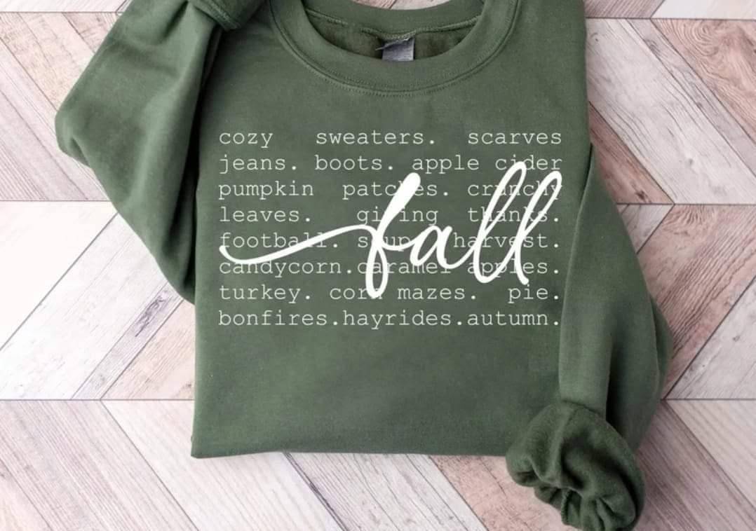 An olive green crewneck sweatshirt with a white typewriter-style text listing of fall items and the word 'fall' in a large white script, laid flat on a light wood herringbone floor.
