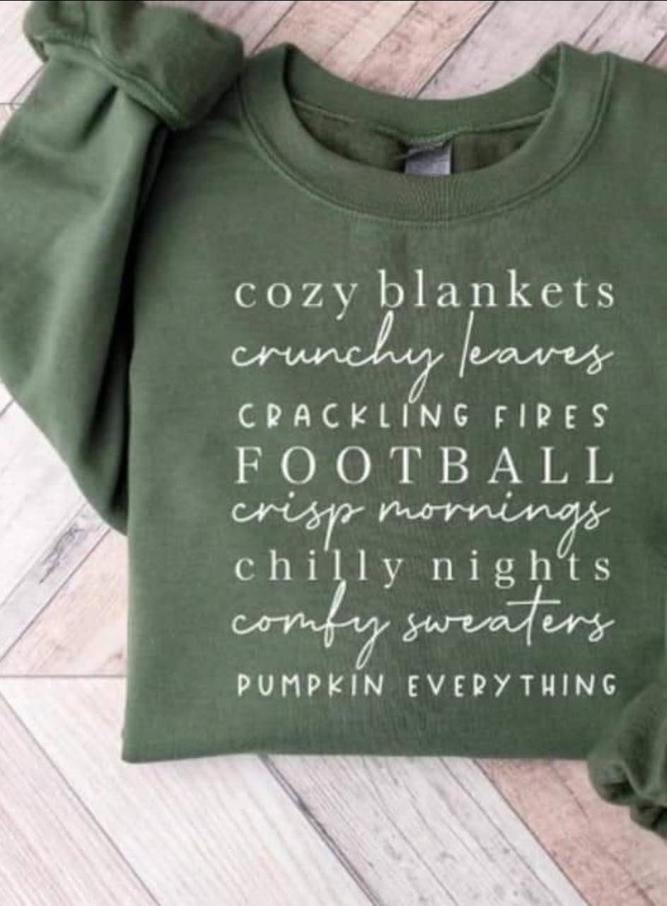 An olive green crewneck sweatshirt with white text listing cozy autumn themes like 'cozy blankets', 'crunchy leaves', 'crackling fires', 'FOOTBALL', 'crisp mornings', 'chilly nights', 'comfy sweaters', and 'PUMPKIN EVERYTHING', laid flat on a light wood plank background.