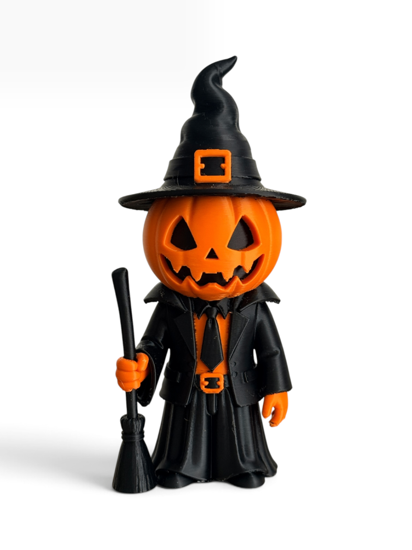 A 3d printed Halloween figurine of a jack-o'-lantern character dressed in a black witch hat, black robe, and black tie, holding a broom. The pumpkin head is bright orange with black facial features, and the hat has an orange buckle detail.