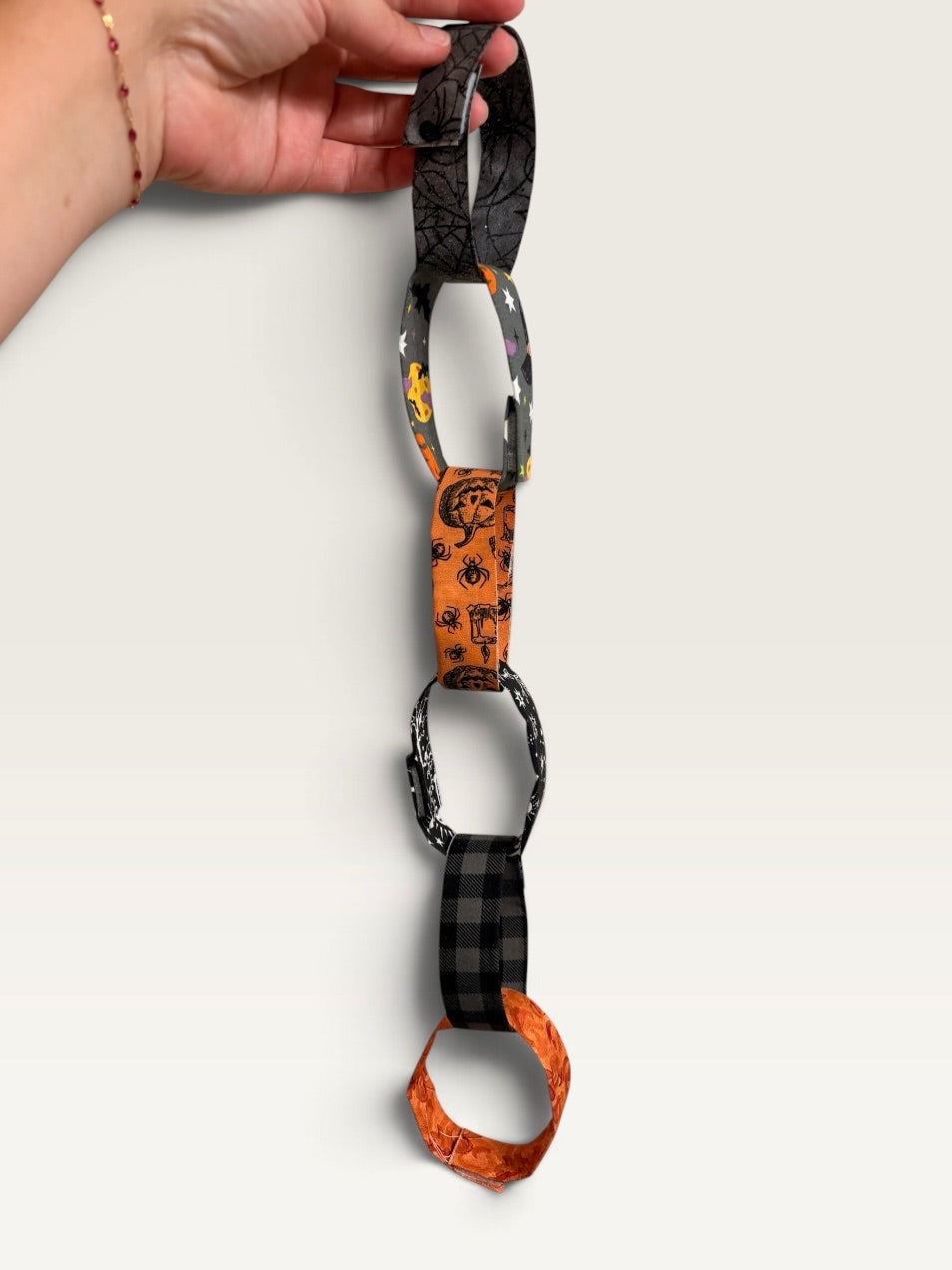A hand holds a handmade paper chain with Halloween-themed patterns, including spiderwebs, skulls, and plaid, against a plain light background.