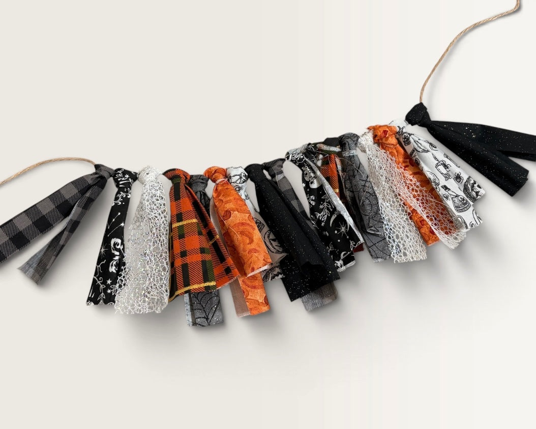 A Halloween-themed fabric garland with multiple tassel-like strips in black, white, orange, and plaid patterns, strung on a natural twine cord against a plain off-white background.