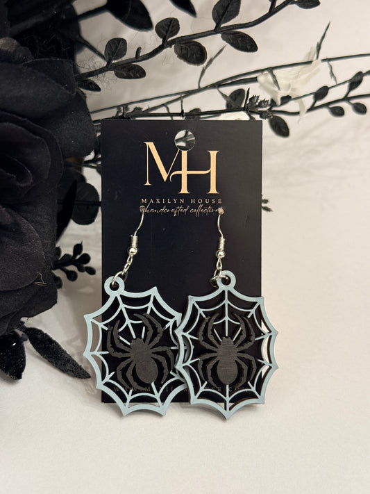 A pair of dangling spiderweb earrings with black spider cutouts and gray tone web outlines, displayed on a black branded card against a light neutral background with decorative black flowers.