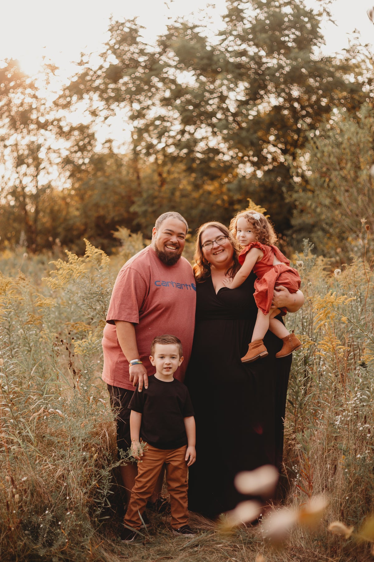 A group of four people, two adults and two children standing looking at the camera smiling in the middle of a grassy field. 