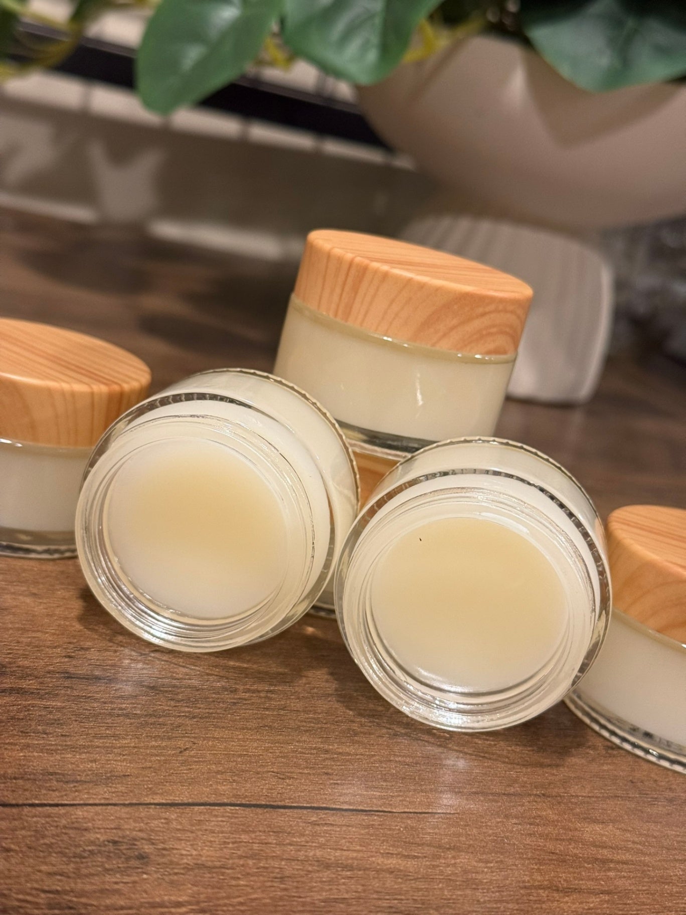 Several small clear cosmetic jars filled with pale cream product, arranged on a wooden surface. Two jars are open and in front, with light wood lids visible; a blurred plant and white planter appear in the background.