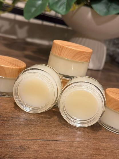 Several small clear cosmetic jars filled with pale cream product, arranged on a wooden surface. Two jars are open and in front, with light wood lids visible; a blurred plant and white planter appear in the background.