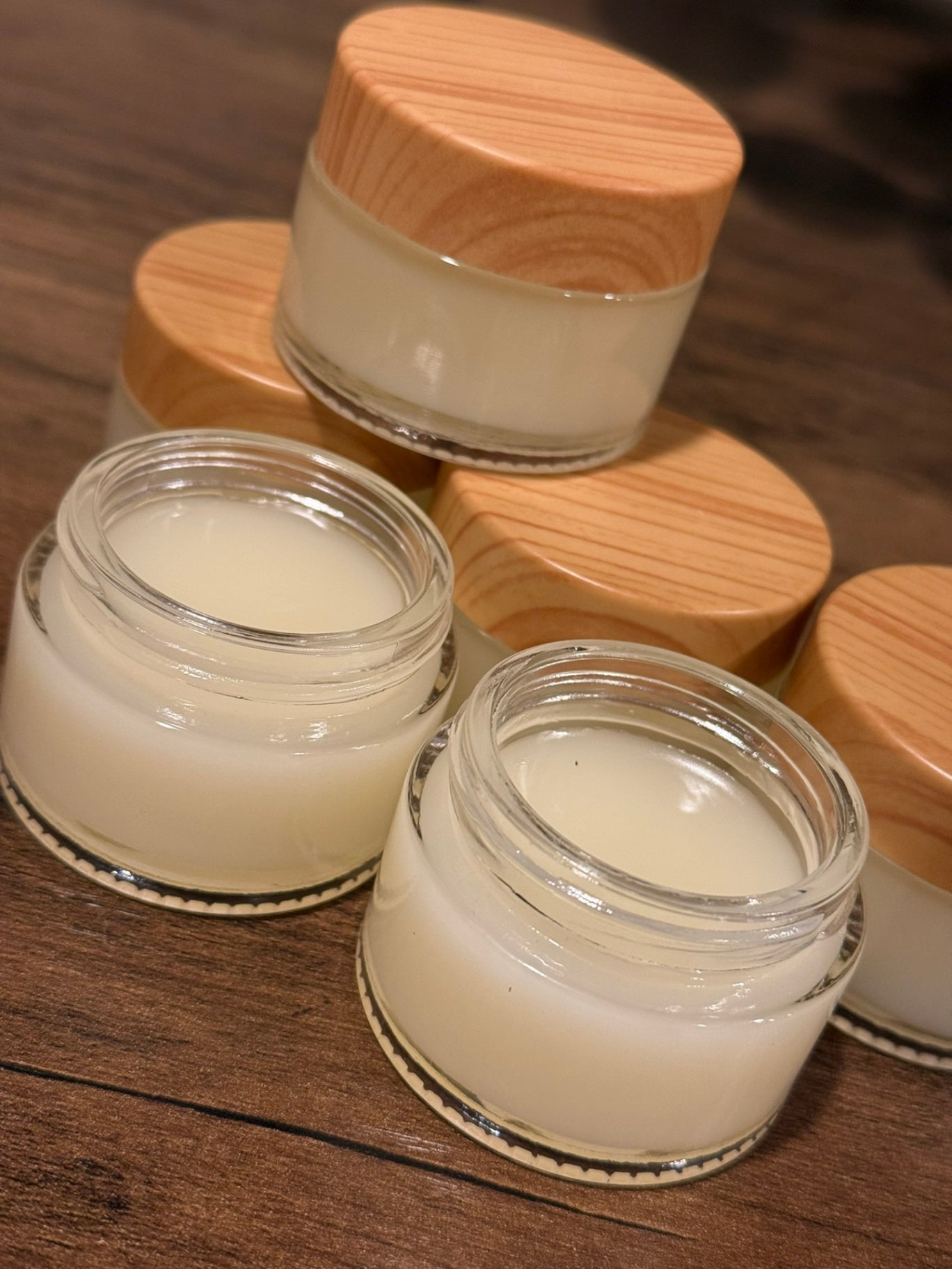 Several small clear cosmetic jars filled with a creamy off-white product, arranged on a dark wooden surface. Three jars have light natural wood lids, and two jars are open.
