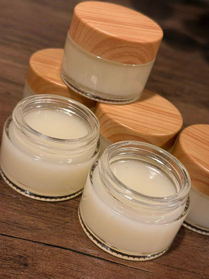 Several small clear cosmetic jars filled with a creamy off-white product, arranged on a dark wooden surface. Three jars have light natural wood lids, and two jars are open.