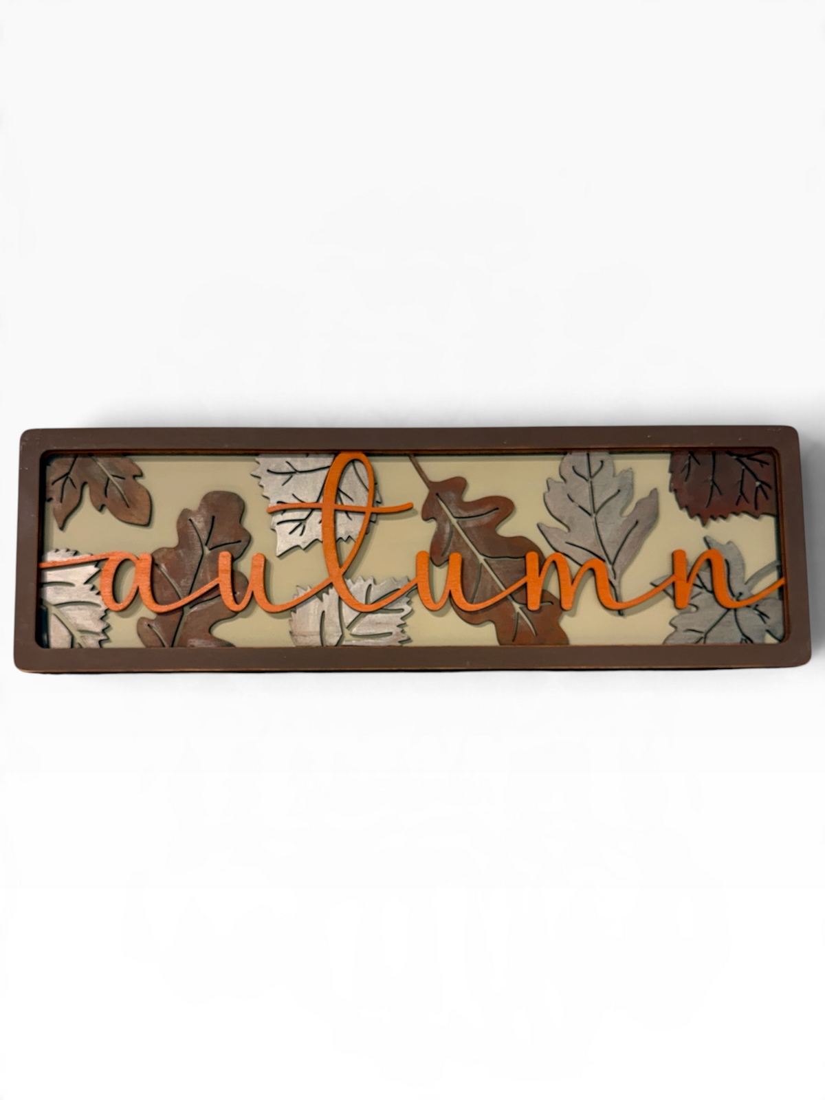 A rectangular wooden sign with the word 'autumn' in orange cursive script layered over a background of layered brown and gray autumn leaves on a cream background, all enclosed in a dark brown frame.