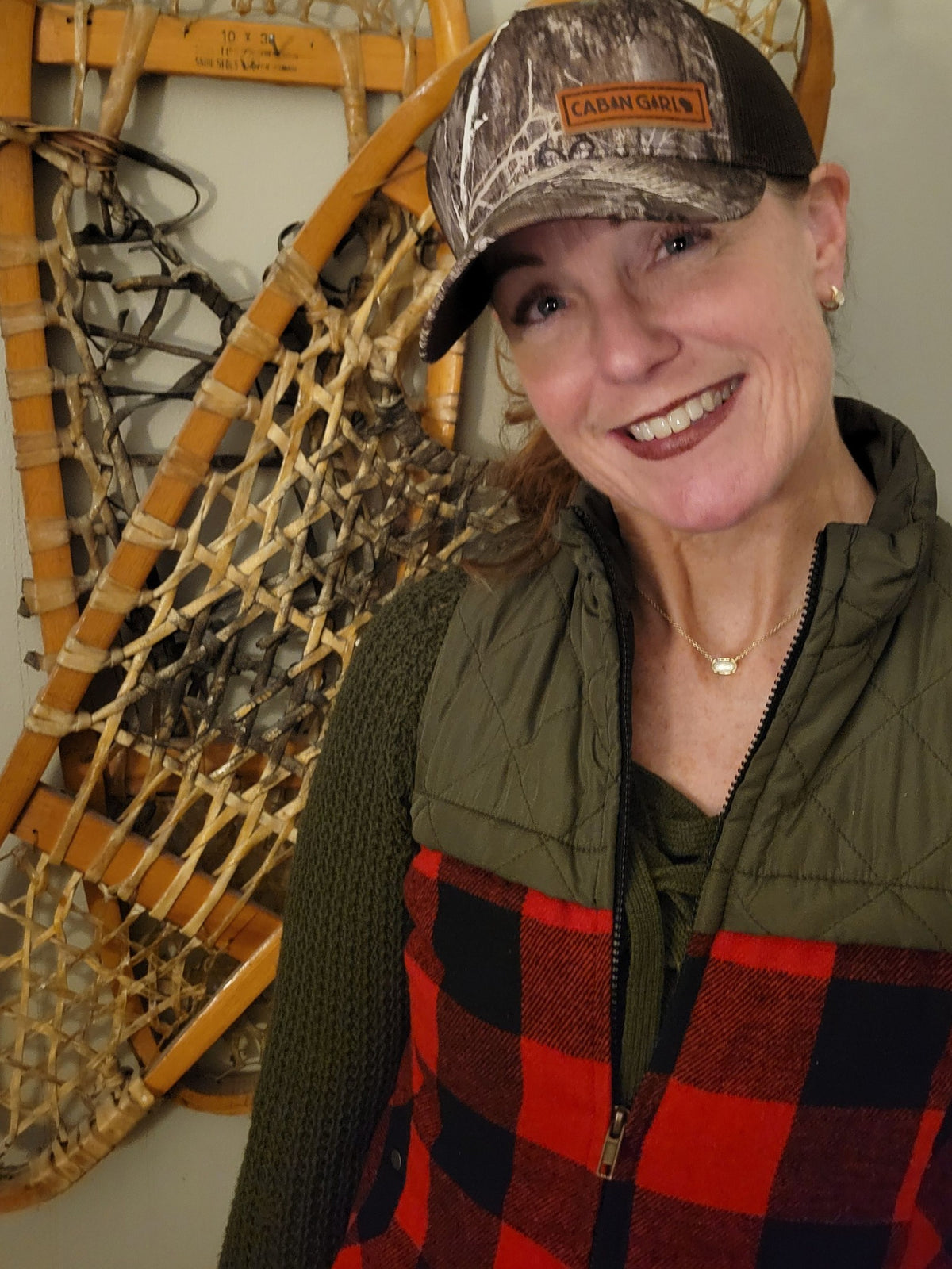 Woman wearing a camouflage cap and green jacket with a red plaid apron, standing next to snowshoes.