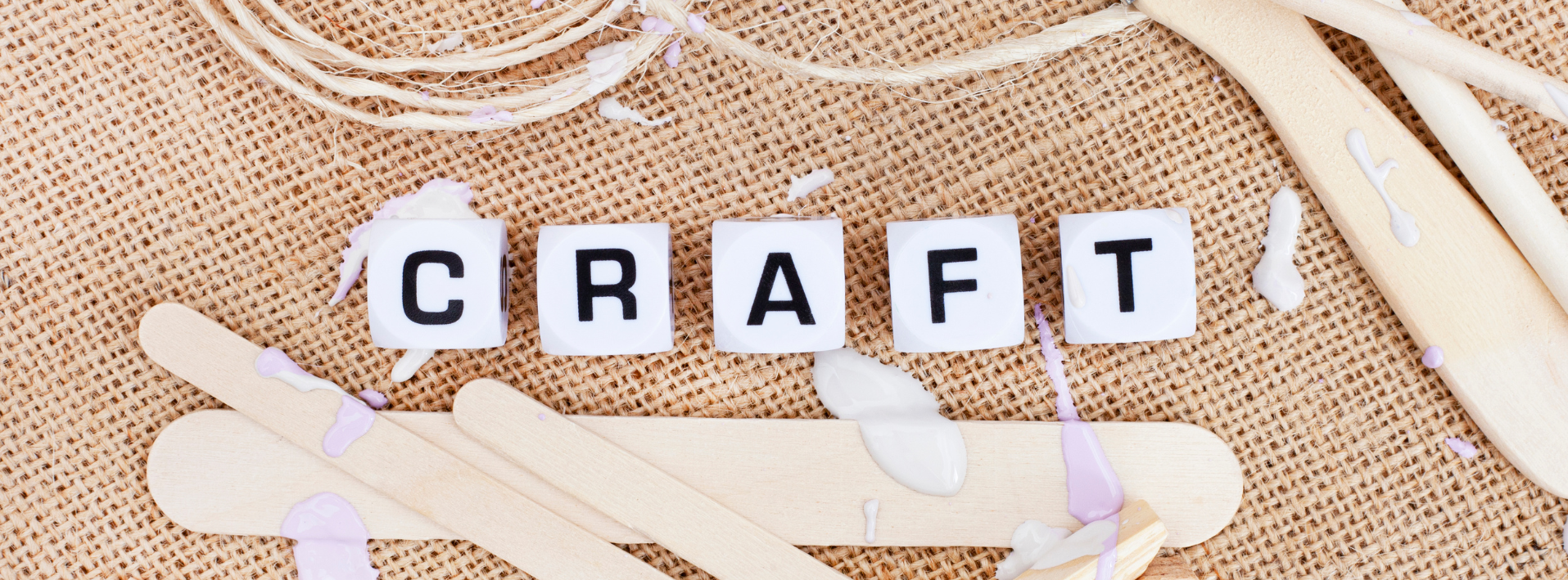 The word 'CRAFT' spelled out with wooden block letters on a textured fabric background with paint sticks, paint, glue splashes.