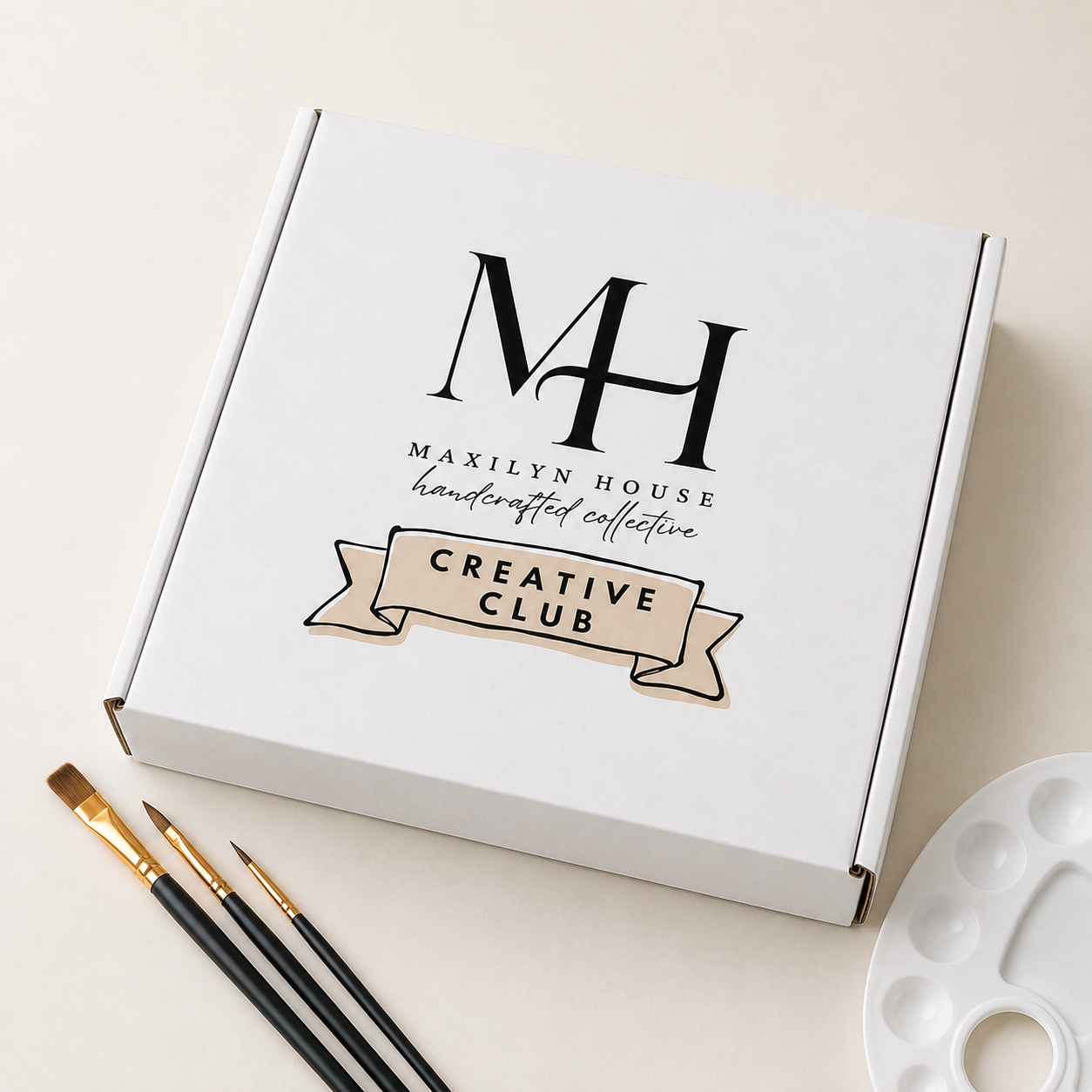 A white square box with the printed logo 'MH' and the text 'Maxilyn House Handcrafted Collective Creative Club' on a light beige surface, styled with paintbrushes and a white paint palette.