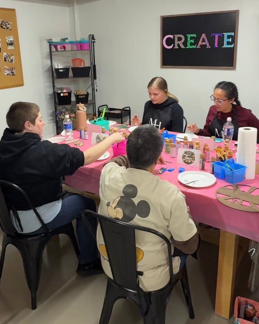 Four people sitting around a table covered in a pink tablecloth, engaged in a painting craft with various supplies like paint bottles, paper, and scissors. A black chalkboard on the wall displays the word 'CREATE' in colorful letters.