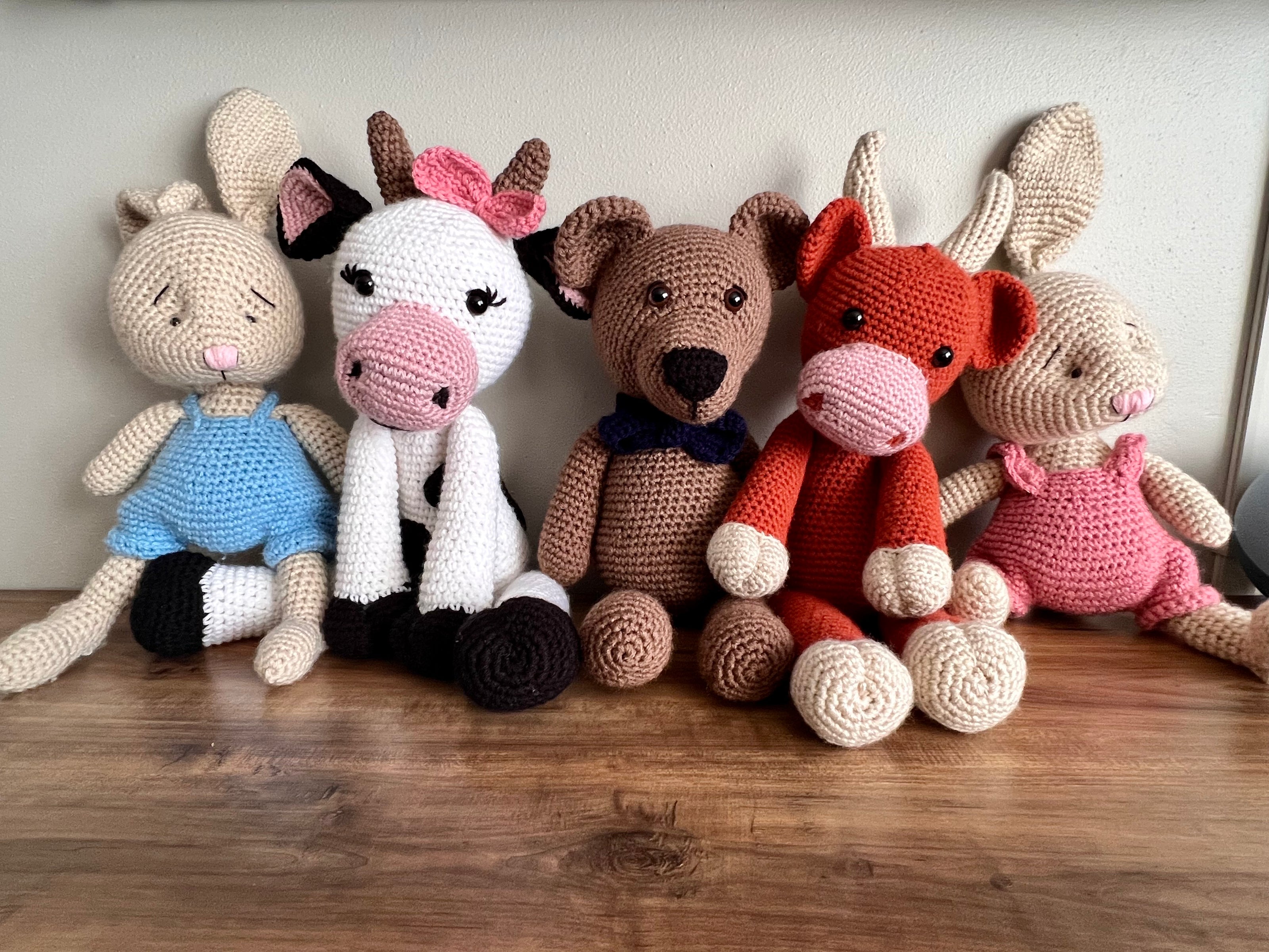 Five different crochet stuff animals propped up next to each other on a wood surface. A cream colored bunny wearing blue overalls. A cow with eyelashes wearing a pink bow on one horn. A teddy bear wearing a blue bowtie. An orange highland cow with corns. A bunny wearing a pink romper. 