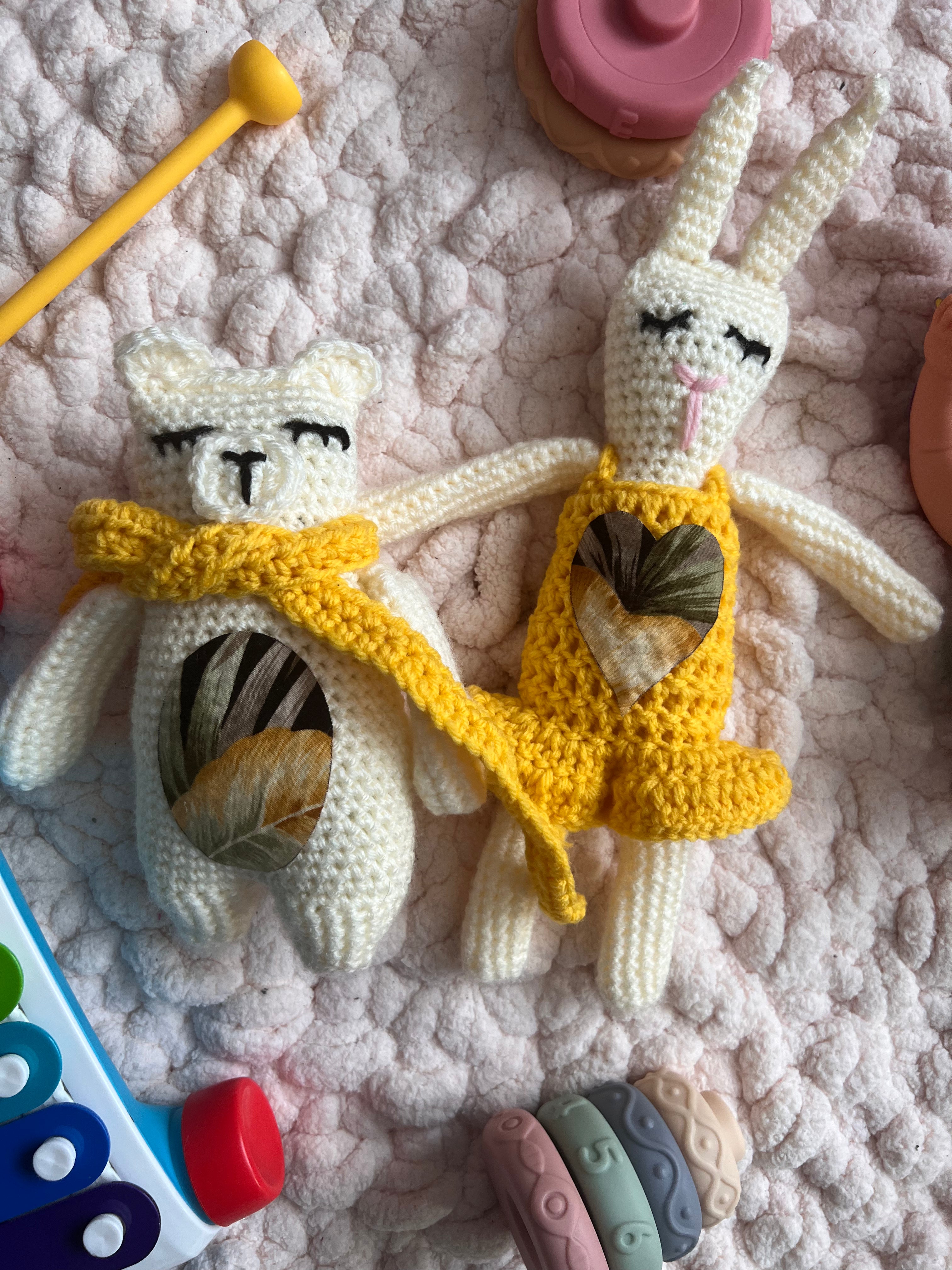 A photo of a crocheted bear and bunny styled on a rug with children's toys around them. The bear has a bright yellow scarf around its neck and the bunny is wearing a yellow crocheted dress. There is a floral patchwork circle on the bears stomach and a patchwork heart on the bunnies dress. 