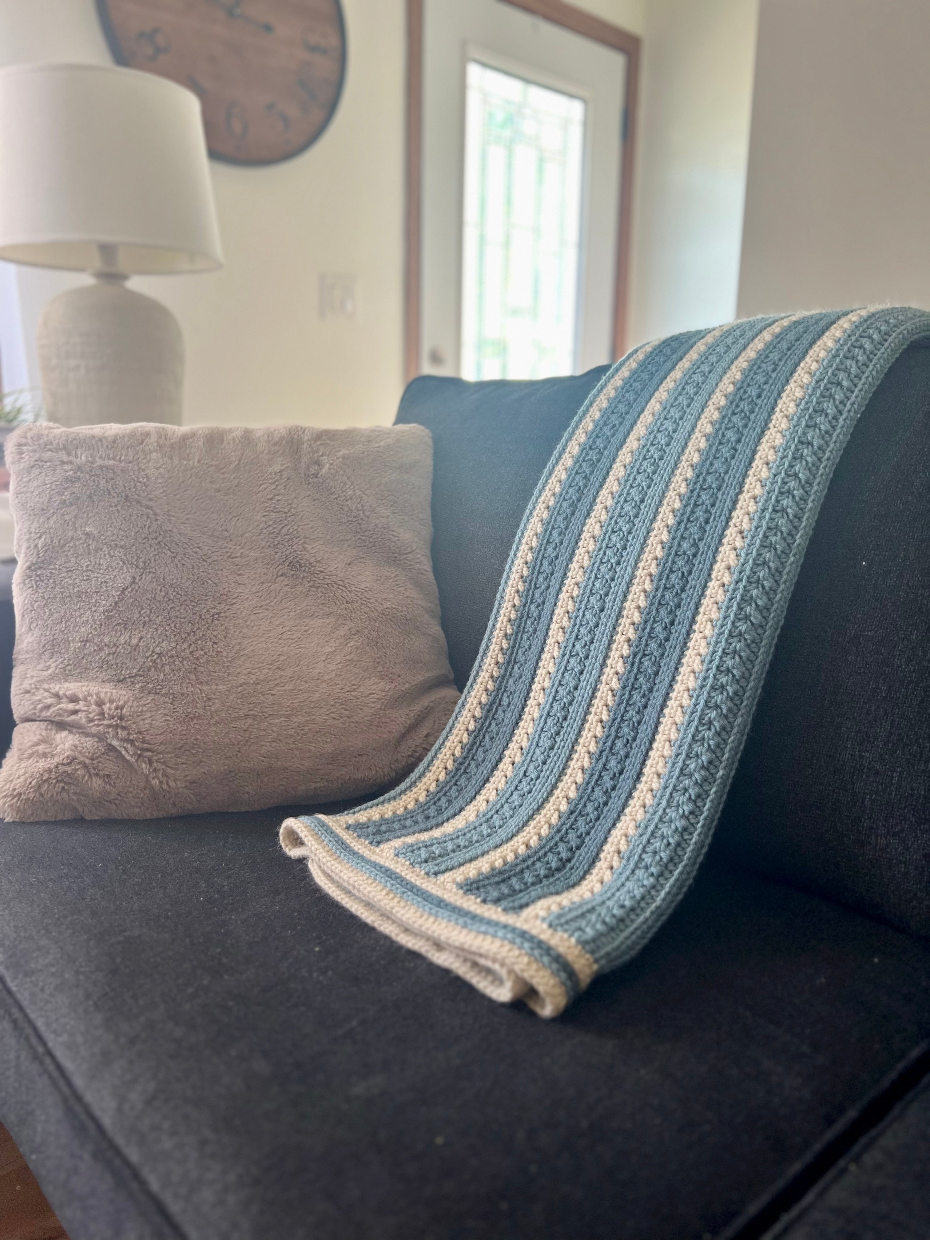 An image of a folded crochet blanket hanging on the back of a dark colored couch. Shows throw pillows, a lamp and a clock in the background.