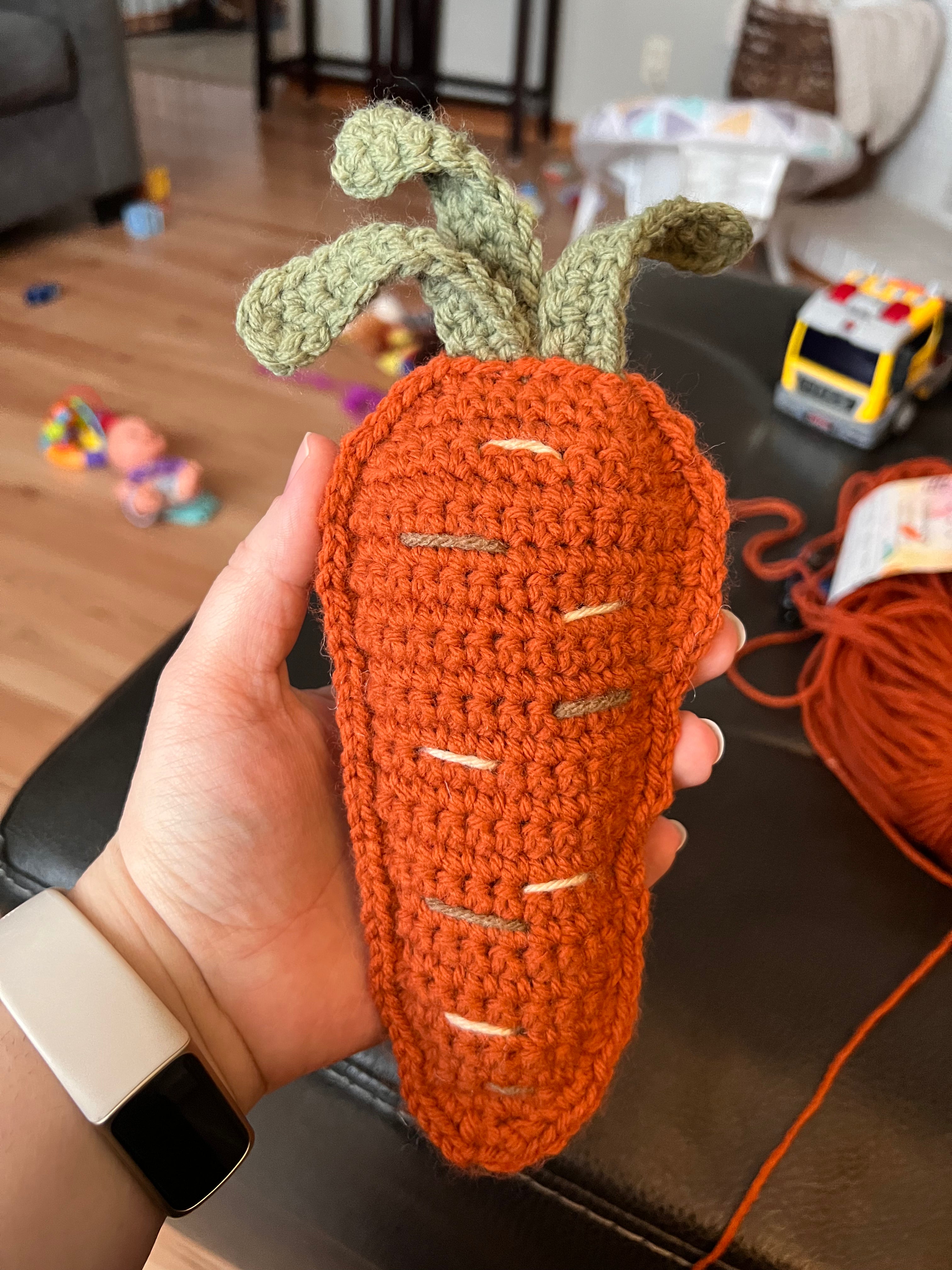 A photo of a crocheted carrot being held by a woman in her hand with a home background behind it.