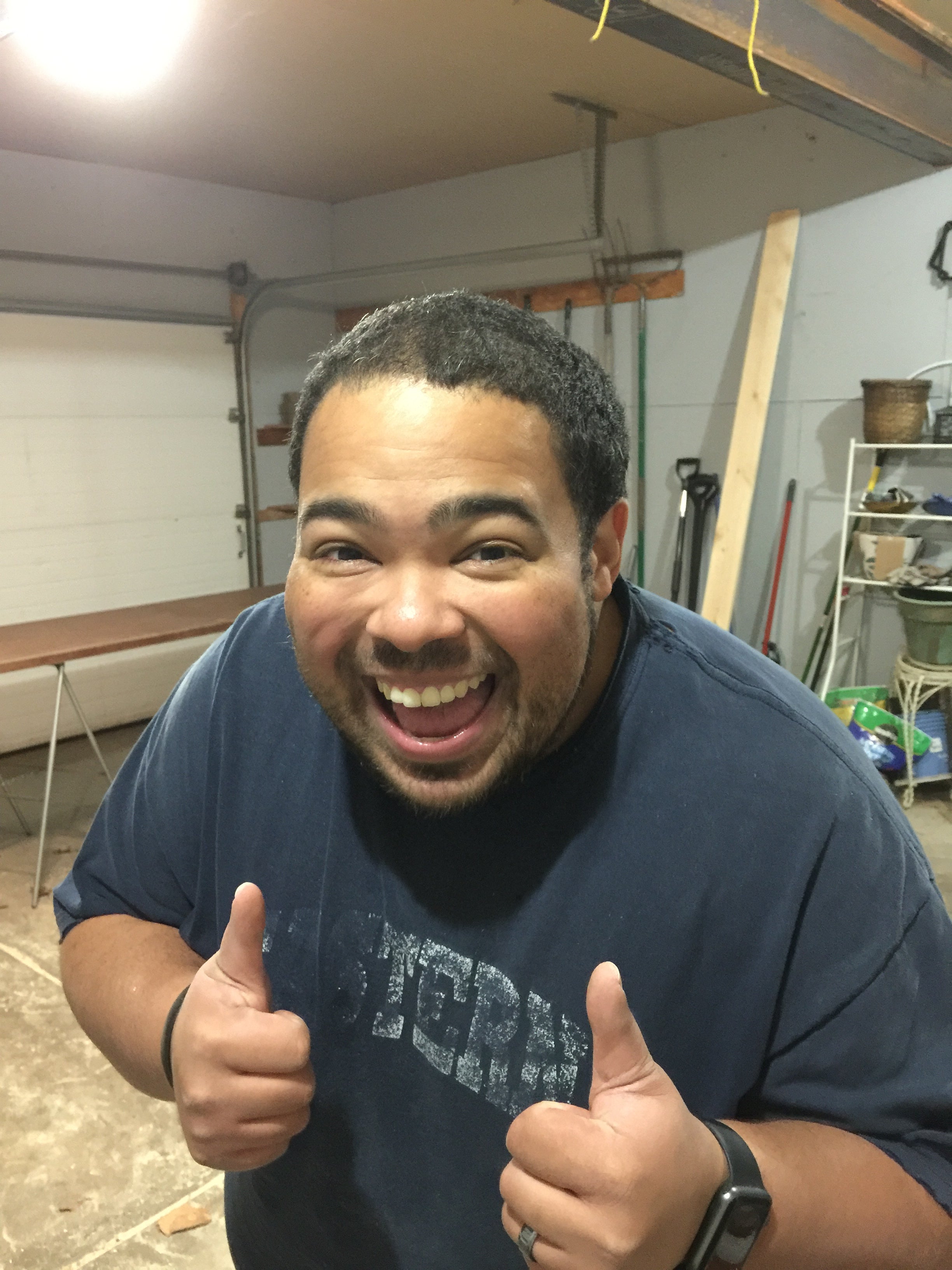 A photo of a man in a garage with tools and other items behind him. He is smiling at the camera and holding two thumbs up.