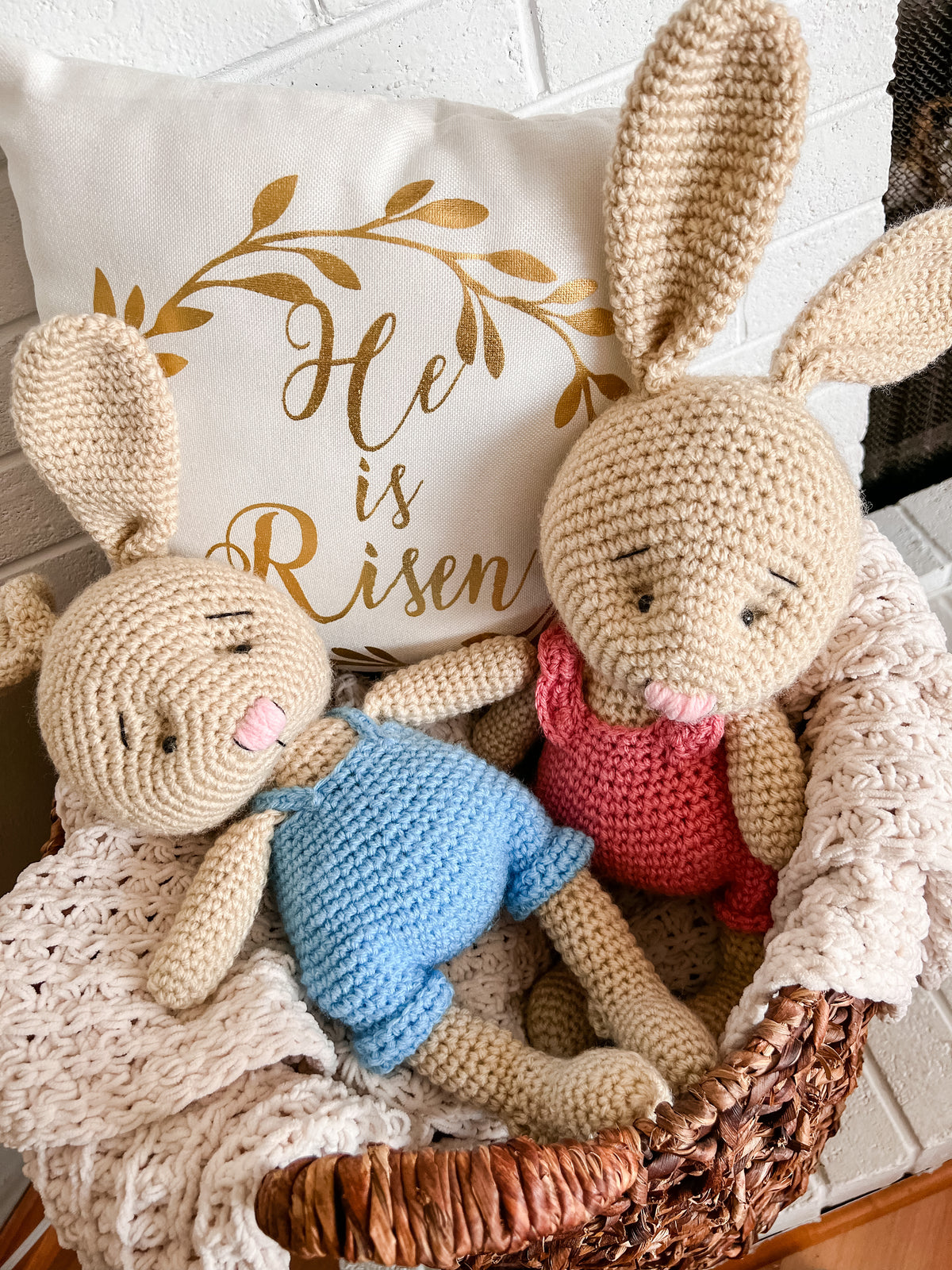 Two crocheted bunnies sitting in a basket with blankets stuffed around it and a pillow reading 'He is Risen' One bunny on the left has blue overalls and the bunny on the right is wearing a pink romper.