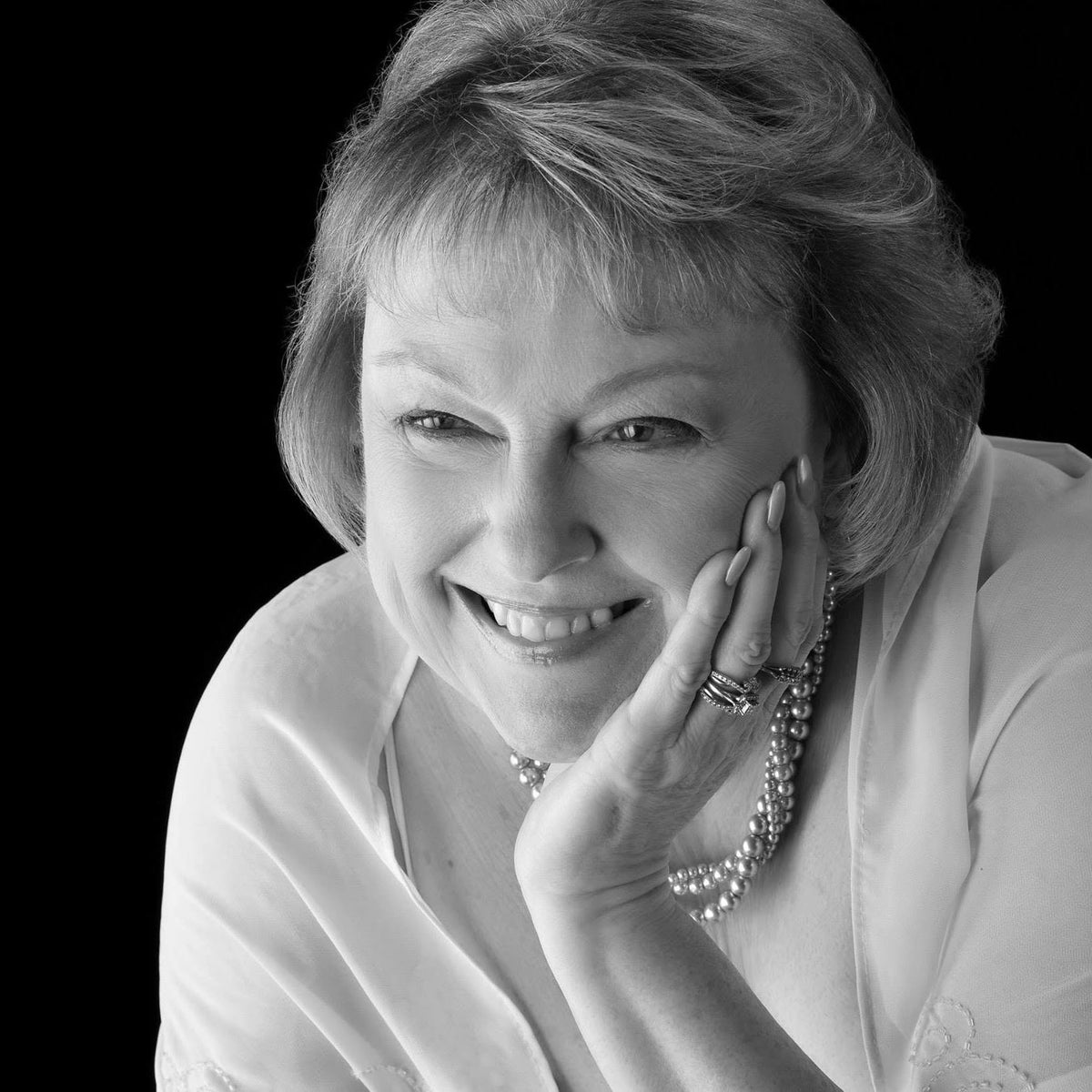 A black and white portrait of a smiling woman with short hair, resting her chin on her hand, wearing a pearl necklace and rings against a solid black background.